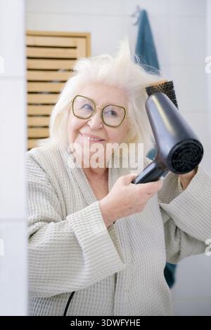 Eine ältere Frau mit weißem Haar und Brille lächelt, während sie im Badezimmer einen Haartrockner benutzt. Sie trägt ein gemütliches Gewand und strahlt Freude und Selbstbewusstsein aus. Stockfoto
