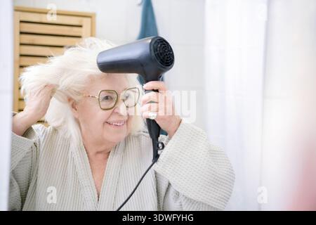 Eine ältere Frau mit Brille und weißem Haar trocknet ihr Haar mit einem schwarzen Haartrockner. Im Bademantel steht sie in einem hellen Badezimmer und lächelt Stockfoto