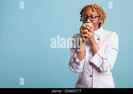 Porträt einer müden Küchenchefin in Brille und Uniform, stehend im Studio mit Kaffeetasse. Überarbeiteter Restaurantarbeiter, der mit geschlossenen Augen das Koffeinaroma genießt und nach der Arbeit einen entspannenden Moment hat. Stockfoto
