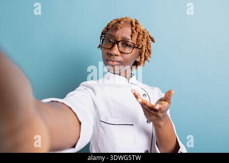 Schwarze Frau in Chefuniform, die täglich Vlog in der Kochschule aufnimmt und Updates mit Online-Zuschauern teilt. Professionelle weibliche Köchin bespricht während eines Videogesprächs am Telefon das Menü für Veranstaltungen mit dem Kunden. Stockfoto