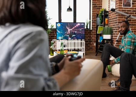 Nahaufnahme einer weißen Frau, die auf der Couch sitzt und sich auf Online-Shooting-Spiele mit Wireless-Controller konzentriert. Junge weibliche Individuen genießen Spielesitzungen im Fernsehen zu Hause mit Freunden. Stockfoto