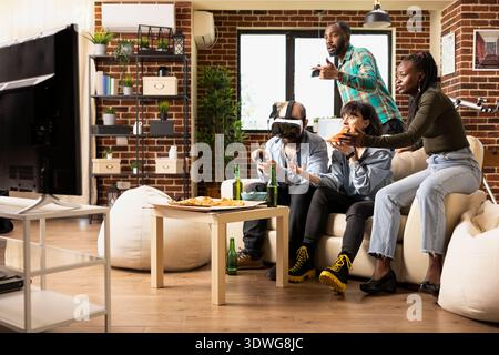 Verschiedene Leute treffen in der Wohnung, reagieren auf kaukasier, der Virtual-Reality-Spiel spielt. Der weiße Mann trägt ein VR-Headset und taucht in interaktive Spiele ein, während Freunde Pizza essen und zuhause zusehen. Stockfoto