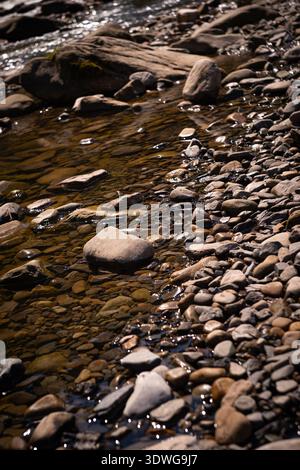 Blick von oben auf einen klaren, flachen Fluss mit glatten Kieseln und Steinen, die durch transparentes Wasser sichtbar sind. Natürlicher Texturhintergrund mit Sonnenlichtreflektio Stockfoto