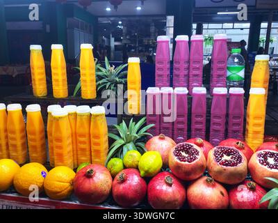 Frische Fruchtsäfte in gelben und rosafarbenen Flaschen werden mit Granatäpfeln und Orangen an einem Marktstand serviert, an dem eine Vielzahl tropischer Früchte präsentiert wird Stockfoto