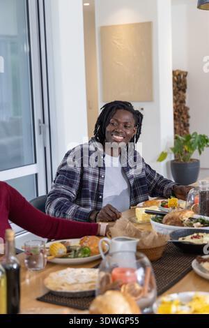 Freunde mit einem Afroamerikaner in kariertem Hemd, der am Esstisch mit Maisbrötchen lächelt Stockfoto