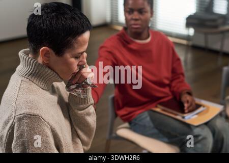 Kaukasische junge Erwachsene Frau, die mit den Handweinen sitzt und verstört erscheint, während schwarze junge Erwachsene Frau in der Nähe sitzt und Unterstützung bei der Gruppentherapie anbietet Stockfoto