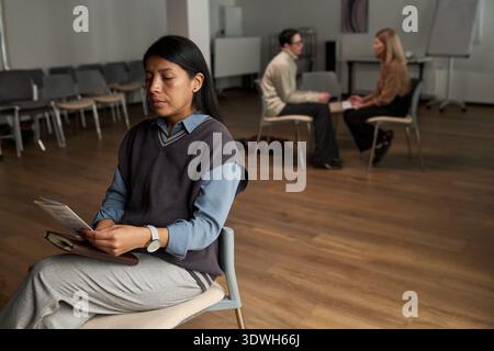 Die hispanische Frau sitzt allein und liest ein Faltblatt im Vordergrund, während zwei kaukasische Frauen im Hintergrund im leeren Besprechungsraum sitzen, Gruppentherapie-Sitzungskonzept Stockfoto