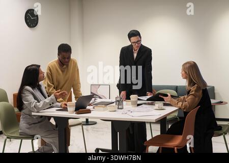 Gruppe von jungen Erwachsenen und Frauen mittleren Alters aus verschiedenen ethnischen Gruppen, die Geschäftsstrategien am Tisch diskutieren, Laptop und Dokumente während eines gemeinsamen Büromeetings verwenden Stockfoto