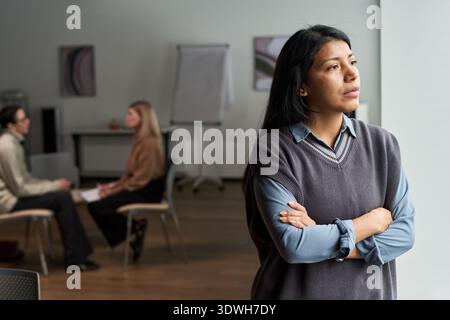 Hispanische Frau, die mit überkreuzten Armen aus dem Fenster steht, zwei kaukasische Erwachsene sitzen im Hintergrund und unterhalten sich im modernen Büro der Psychologen Stockfoto