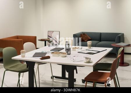 Moderner Büroarbeitsplatz mit leerem Tisch mit verstreuten Dokumenten, Kaffeetassen, digitalen Tablets, Schreibwaren und Stühlen, Hintergrund mit Sofabühlen und minimalistischem Dekor Stockfoto