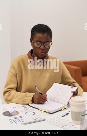 Kaukasische junge Erwachsene Frau, die eine Brille trägt, am Schreibtisch sitzt, Finanzpläne analysiert und Notizen im Planer schreibt, einen Stift mit einer Kaffeetasse und Dokumente auf dem Tisch hält Stockfoto