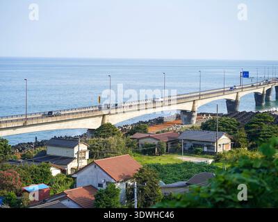 Ein Blick aus einem hohen Winkel auf eine lange Betonstraße, die sich über das blaue Meer erstreckt, und ein üppiges Küstendorf in Hitachi Japan Stockfoto