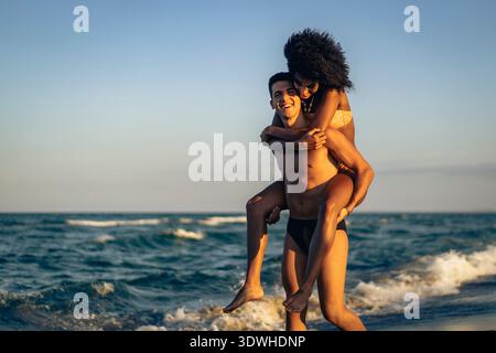 Glückliches, junges, multiethnisches Paar, das zur goldenen Stunde einen verspielten Huckepack-Ritt im Meer macht, um Liebe und Sommerspaß zu feiern. Stockfoto