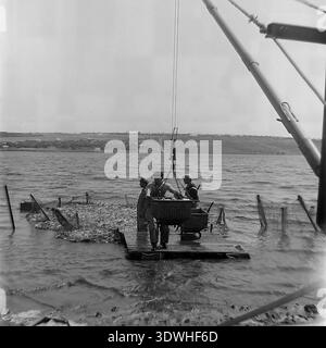 Das historische Schwarzweiß-Foto aus den 1970er Jahren zeigt ein Team von drei Fischern auf einem Holzfloß, das einen riesigen Fischfang aus einem Teich im Donrybkombinat im Bezirk Slavjansk, ukrainische SSR, UdSSR, zieht. Sie verwenden ein großes Netz und einen mechanischen Kran, um einen schweren Korb mit frischem Fisch zu heben. Dieses authentische Archivbild zeigt die industrielle Dimension der sowjetischen Aquakultur, harte Teamarbeit und die Entwicklung der Lebensmittelindustrie im friedlichen Donbass vor dem Krieg, was den landwirtschaftlichen Wohlstand widerspiegelt. Stockfoto