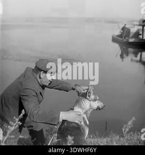 Das historische Schwarzweißfoto aus den 1970er Jahren zeigt einen sowjetischen Fischer, der auf dem Grasufer eines Teichs ruht und seinen Hund sanft im Donrybkombinat im Bezirk Slawjansk, ukrainische SSR, UdSSR streichelt. Er trägt eine traditionelle flache Kappe und hält eine Zigarette und teilt einen ruhigen Moment mit dem Tier, während ein motorisiertes Boot im Hintergrund gleitet. Dieses authentische Archivbild zeigt das tägliche landwirtschaftliche Leben, die Mensch-Tier-Gemeinschaft und den friedlichen Donbass vor dem Krieg, was eine ruhige ländliche Atmosphäre widerspiegelt. Stockfoto