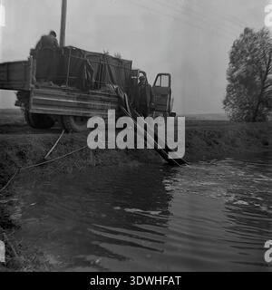 Das historische Schwarzweißfoto der 1970er Jahre zeigt sowjetische Arbeiter, die einen Teich im Donrybkombinat im Bezirk Slawjansk, ukrainische SSR, UdSSR, bestückt haben. Ein spezieller Lkw, der auf einem Damm geparkt wird, verwendet eine lange, flexible Rutsche, um kleine Fische ins Wasser zu bringen. Dieses authentische Archivbild zeigt die technische Ausrüstung der sowjetischen Aquakultur, die landwirtschaftliche Instandhaltung und die stetige Entwicklung der Lebensmittelindustrie im friedlichen Donbass vor dem Krieg, was die industrielle Macht und die organisierte sozialistische Arbeit der Region widerspiegelt. Stockfoto