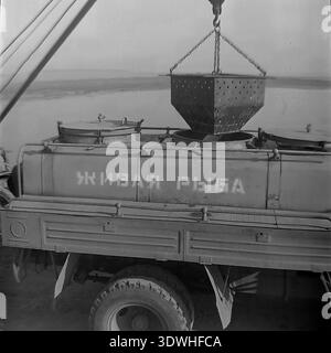 Das historische Schwarzweißfoto der 1970er Jahre zeigt einen Kran, der eine große perforierte Metallkiste mit frischem Fisch in einen speziellen Transportwagen mit der Aufschrift „Live Fish“ (Живая рыба) im Donrybkombinat im Bezirk Slavjansk, ukrainische SSR, UdSSR, lädt. Dieses authentische Archivbild zeigt die technische Mechanisierung, die effiziente Logistik der Lebensmittelindustrie und die stetige Entwicklung der Aquakultur im friedlichen Donbass vor dem Krieg. Stockfoto