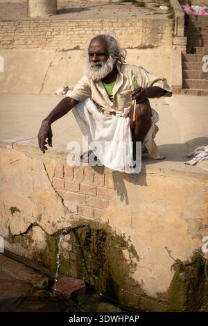 Ein älterer Mann putzt sich die Zähne in der Nähe eines Wasserauslaufs in Varanasi, Indien, und zeigt das tägliche Leben und die Tradition. Stockfoto