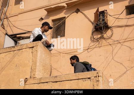 Zwei Männer sitzen und unterhalten sich auf einem Dach vor einem beigefarbenen Gebäude in Varanasi, Indien, mit sichtbaren elektrischen Leitungen. Stockfoto