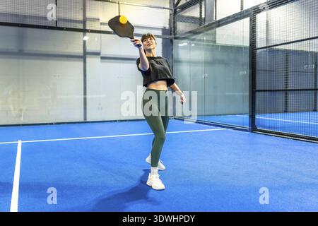 Frau in sportlicher Kleidung spielt Pickleball auf einem leuchtend blauen Hallenplatz, schwingt ein Paddel, um den gelben Ball zu treffen und genießt aktiven Sport und Passform Stockfoto