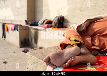 Zwei Menschen ruhen sich auf Steinbänken in Varanasi, Indien aus, mit Kleidung und Taschen neben ihnen im Sonnenlicht. Stockfoto