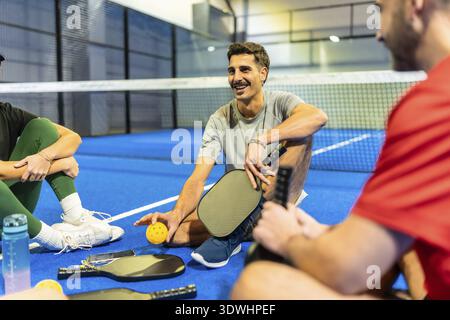 Verschiedene Gruppen junger erwachsener Freunde, die nach dem Spielen mit ihren Paddeln und Ba einen Moment der Ruhe und Unterhaltung auf einem blauen Pickleball-Platz genießen Stockfoto