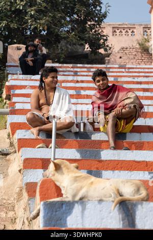 Zwei junge Männer entspannen sich auf gemalten Stufen mit einem Hund in Varanasi, Indien, und genießen einen sonnigen Tag im Freien. Stockfoto