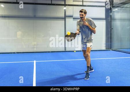 Ein Mann spielt Padel auf einem leuchtend blauen Platz, hält einen Paddelschläger und trifft während eines intensiven Spiels einen hellgelben Pickleball, der Action A zeigt Stockfoto