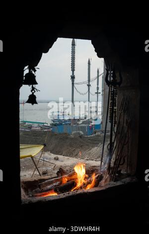 Heiliges Bestattungsfeuer brennt am Ufer des Ganges in Varanasi, Indien, mit Blick auf die Boote und den Fluss im Hintergrund. Stockfoto