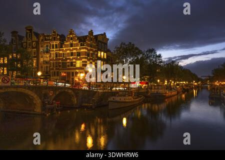 Niederlande. Wolkiger Abend auf dem Amsterdamer Kanal. Boote und Hausboote sind vertäut. Reflexion im Wasser der traditionellen Häuser und einer Brücke Stockfoto