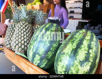 Wassermelonen und Ananas sind in einer Holzkiste an einem Saftstand angeordnet, während eine Frau im Hintergrund Getränke zubereitet Stockfoto