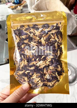 Getrocknete Schmetterlingserbsenblüten werden in einem transparenten Goldpaket dargestellt, das von einer Hand gehalten wird, wobei eine Kücheneinrichtung im Hintergrund sichtbar ist Stockfoto