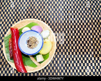 Frische Zutaten und Gewürze für die thailändische Küche auf einem Bambusteller, einschließlich Chili, Kräuter und einer Schüssel mit Gewürzen auf einer gewebten Matte Stockfoto