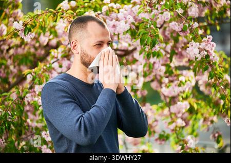 Allergie des Mannes mit saisonaler Allergie im Frühjahr im blühenden Garten im Frühling. Mann niest und bläst Nase mit Nasentaschentuch vor blühendem Baum. Frühjahrsallergiekonzept. Stockfoto