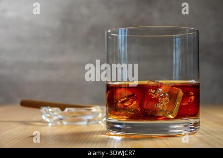 Glas Whiskey mit Eiswürfeln auf dem Holztisch. Ein Aschenbecher mit einer Zigarre im Hintergrund in Unschärfe-Effekte Stockfoto