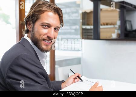 Geschäftsmann im dunklen Anzug, der am Kaffeetisch neben dem Fenster Papier schreibt, hält Stift, Tafel-Menü. Executive, Professional, Kaffeehaus, Sonnenlicht, Dokume Stockfoto