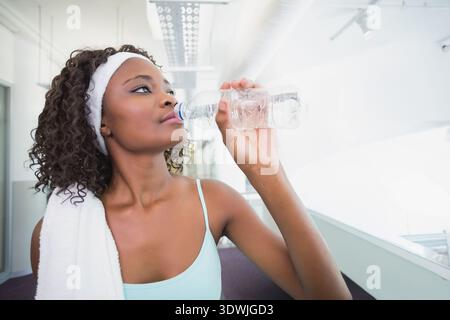 Erwachsene afroamerikanische Frau, die im Fitnessstudio pausiert, aus einer durchsichtigen Flasche trinkt, weißem Handtuch, blauem Tank. Athlet, Fitness, Flur, Atrium, Glas, Geländer, m Stockfoto