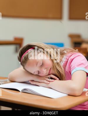 Grundmädchen, das auf gefalteten Armen am Schreibtisch im Klassenzimmer mit offenem Notizbuch, Bleistift und rosa Stirnband ruht. Schüler, Ruhe, Lernen, Ruhe, Konzentration, Holz, quie Stockfoto