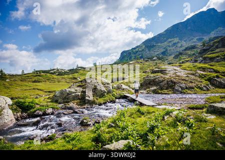 Wanderer überquert eine kleine Holzbrücke über rauschende Gebirgsbach. Malerische Alpenlandschaft in der Steiermark, Österreich mit grünen Hängen und hohen Gipfeln Stockfoto