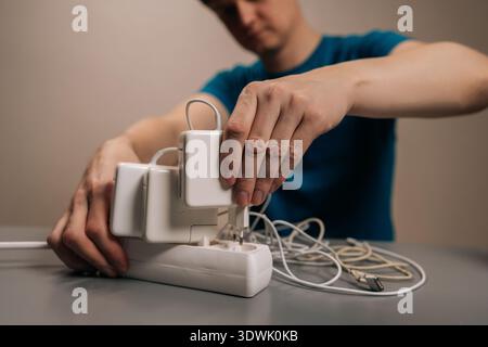 Zugeschnittene Aufnahme von Menschenhänden, die ein rechteckiges weißes Netzteil zum Laden eines elektronischen Geräts, eines Laptops, mit einer organisierten elektrischen Verlängerung verbinden Stockfoto