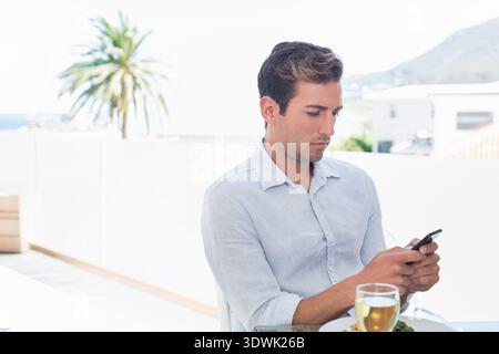Mann in blauem Hemd, der auf der Terrasse sitzt und ein Smartphone hält, Wein trinkt und Salat isst, Kopierraum. Balkon, Küste, Außenbereich, Sonnenlicht, Palme, Tabelle, DE Stockfoto