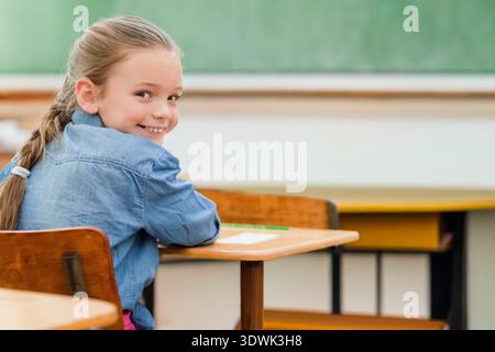 Schülerin, die am Holzschreibtisch sitzt, dreht sich, lächelt mit Papier und grünem Bleistift. Klassenzimmer, Tafel, Schreibtische, Schüler, Mädchen, Blick, l Stockfoto