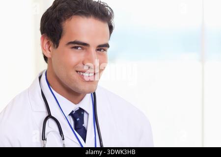 Männlicher Arzt lächelnd und stehend in der Arztpraxis, mit weißem Labormantel und Stethoskop. Arzt, Klinik, Profi, Porträt, Highkey, Mindestwerte Stockfoto