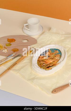 Kreativer Arbeitsbereich mit einer Farbpalette mit Farben, Pinseln und gerahmtem Bildmaterial auf einer hellen Oberfläche, mit einer Tasse im Hintergrund Stockfoto