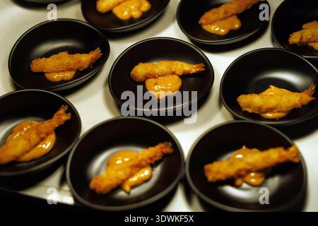 Mehrere kleine schwarze Keramikschalen enthalten jeweils einen knusprigen, panierten Garnelenschwanz, der auf einem Klecks Orangenrahmsauce ruht. Die Vorspeisen sind arra Stockfoto