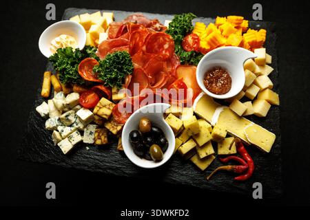 Eine Platte aus dunklem Schiefer wird mit Salami, Schinken, Käsewürfeln, Oliven und kleinen Schüsseln Dip belegt. Frisches Gemüse wird als Garnitur verwendet. Stockfoto