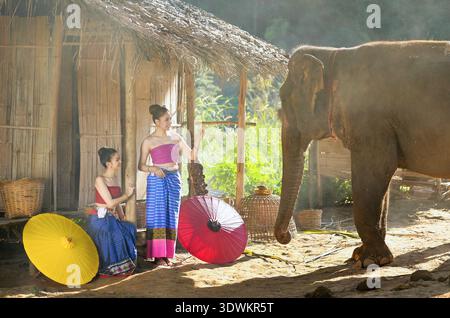 Seitenansicht eines Elefanten, der vor zwei Frauen vor einer Hütte in traditioneller thailändischer lanna-Kleidung steht, Chiang Mai, Thailand Stockfoto