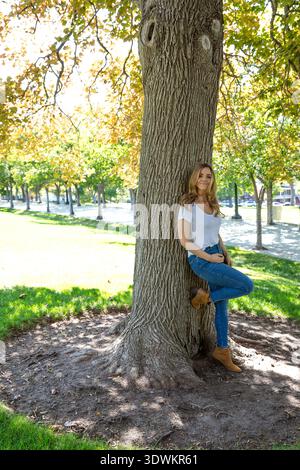 Porträt einer lächelnden Frau in T-Shirt und Jeans, die in einem Park steht und sich mit erhobenem Knie an einen Baum lehnt Stockfoto