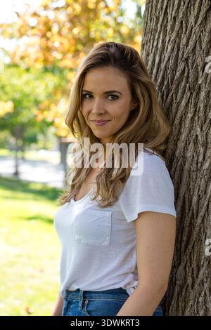 Porträt einer schönen Frau in T-Shirt und Jeans, die in einem Park steht und sich an einen Baum lehnt Stockfoto