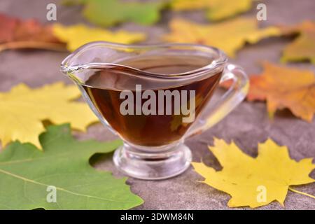 Süßer Ahornsirup in einem Glas Soße Boot und Ahornblätter auf grauem Hintergrund. Stockfoto
